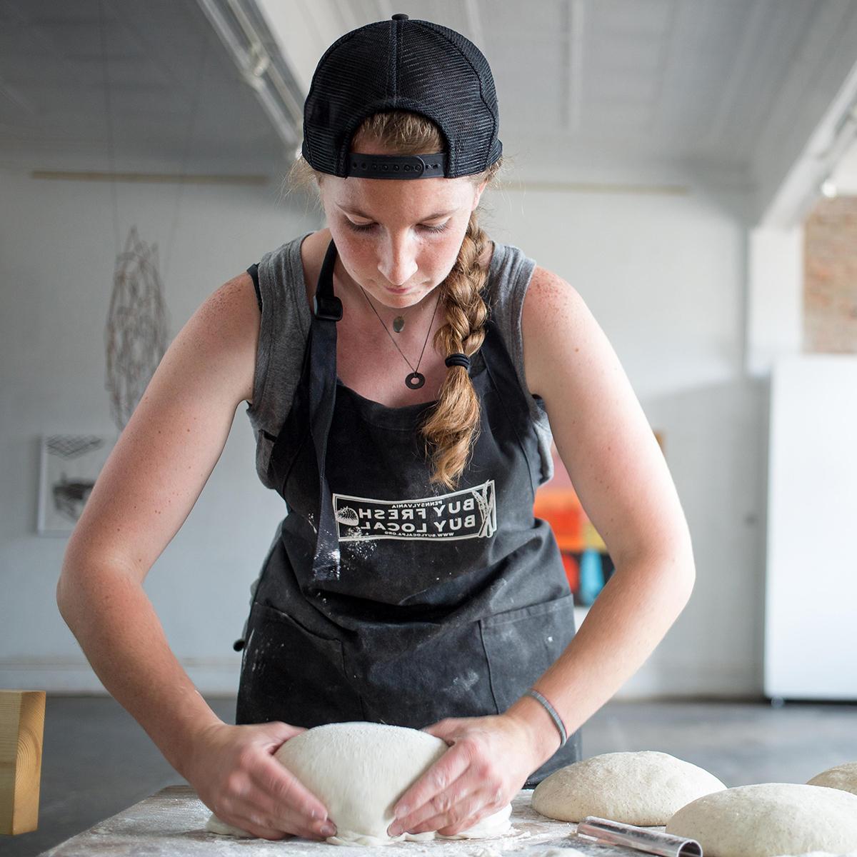 Photo of a young white woman with a black hat, worn backwards, a long braid is kneading dough on a table